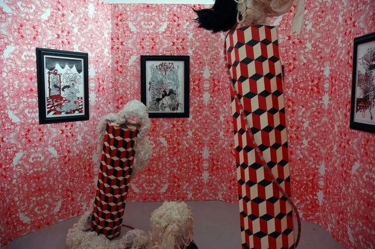 Exhibition room with patterned red wallpaper, framed artworks and sculptures